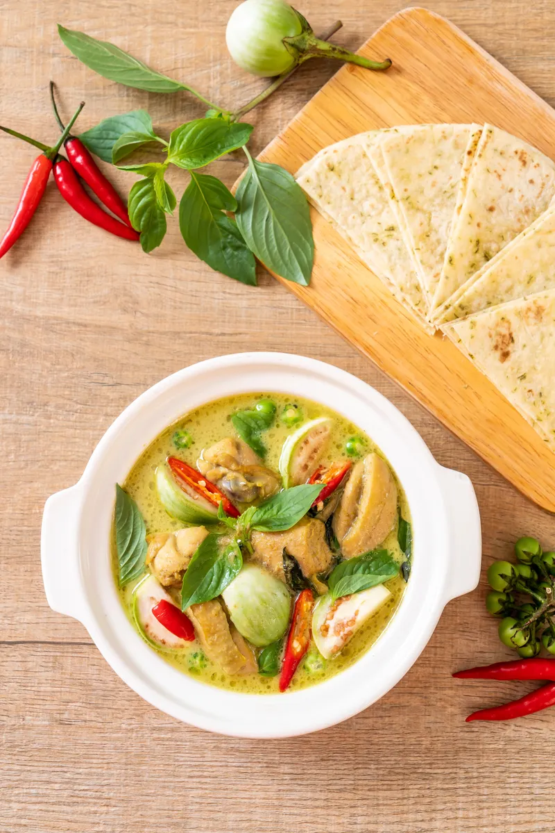 Sumptuous green curry in a whte bowl, garnished with fresh Thai basil and vibrant red chili slices, set against a rustic wooden background.